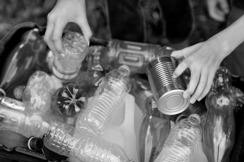 A person is seen sorting and handling discarded plastic bottles and aluminum cans, placing them into a collection container or a bag for waste removal. The plastic bottles vary in size and shape, with some having ridged surfaces and others being smooth, made of transparent or semi-transparent plastic. The aluminum cans are cylindrical, with one clearly showing a metallic, slightly reflective finish, typical of drink cans. The scene suggests an outdoor environment, possibly a recycling or rubbish collection process, with the person's hands actively organizing the waste. The background is blurred, focusing attention on the waste and the handling action. This image illustrates a routine task of separating recyclable waste materials, aligning with objectives of professional rubbish removal services such as those offered by Commercial Waste Parsons Green, to responsibly manage waste disposal in the Parsons Green area.