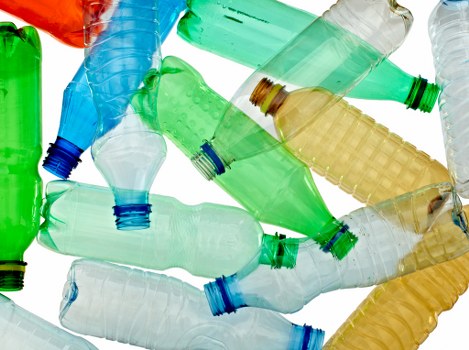 A collection of crushed plastic bottles in various colours, including green, blue, clear, and amber, scattered in a pile. The bottles are made of PET plastic with textured surfaces, some with visible ridges and embossed details. They are positioned in different orientations, with open necks facing upward or sideways, and a few partially crushed or flattened. The scene is set against a plain white background, highlighting the translucency and vibrant tones of the bottles. This assortment of recyclable plastics reflects typical waste items handled during rubbish removal in Parsons Green, London, by companies such as Commercial Waste Parsons Green, and illustrates common types of waste collected in local waste management services.