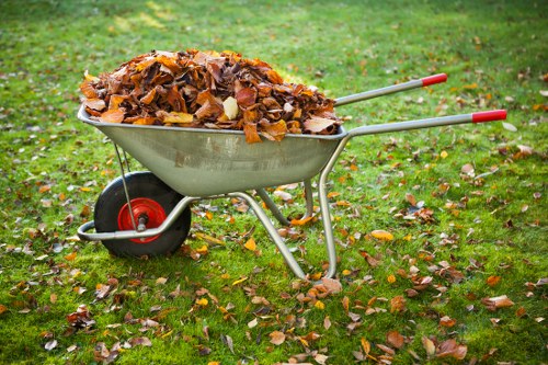 A metal wheelbarrow filled with a large heap of fallen autumn leaves in shades of brown, yellow, and orange, resting on a grassy outdoor area with scattered leaves on the ground. The wheelbarrow has red plastic handles and a black rubber wheel, positioned on a patch of green grass that appears slightly damp. The background shows additional grass with more fallen leaves, suggesting a garden or park setting. The scene is well-lit with natural daylight, highlighting the textures of the leaves and the metallic surface of the wheelbarrow. This image relates to outdoor waste clearance, typical of rubbish removal services offered by Commercial Waste Parsons Green in the Parsons Green area, providing visual context for clearing garden debris and autumn leaf disposal.