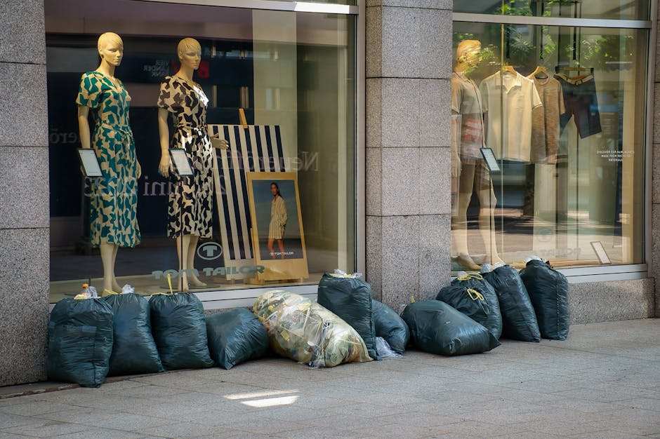 A black wheelie bin labeled 'St. John's' positioned on a sidewalk curb at night, filled with various waste items including cardboard boxes and plastic bags, with the lid partially open. The bin is sit