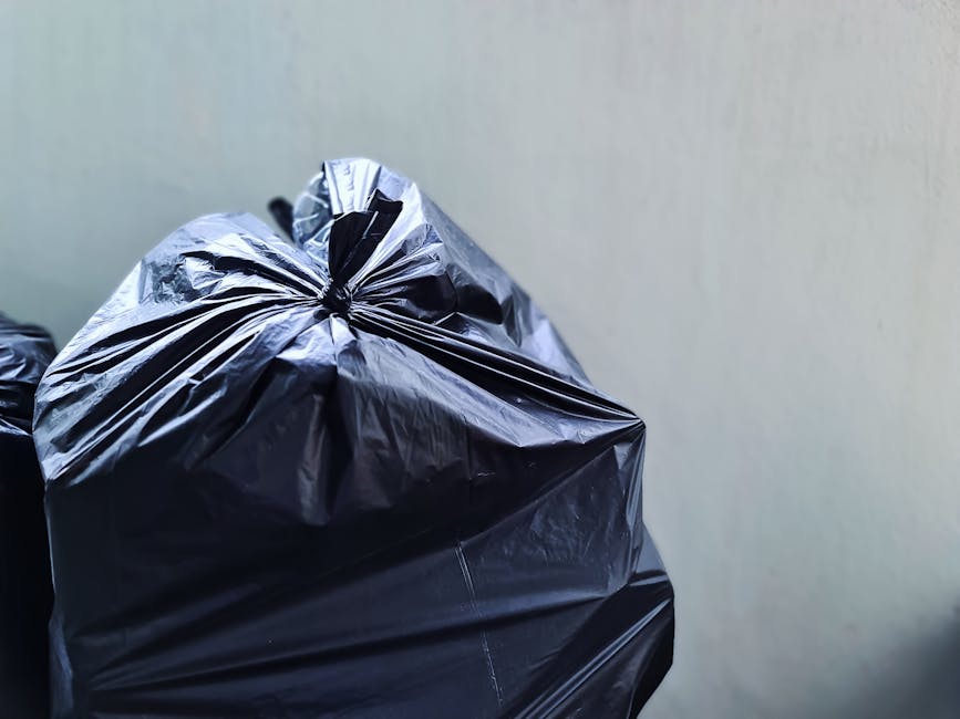 A clear plastic bag filled with crushed or flattened transparent plastic bottles, held by a person wearing a yellow garment. The plastic bottles show visible ridges, caps, and indentations, with some 