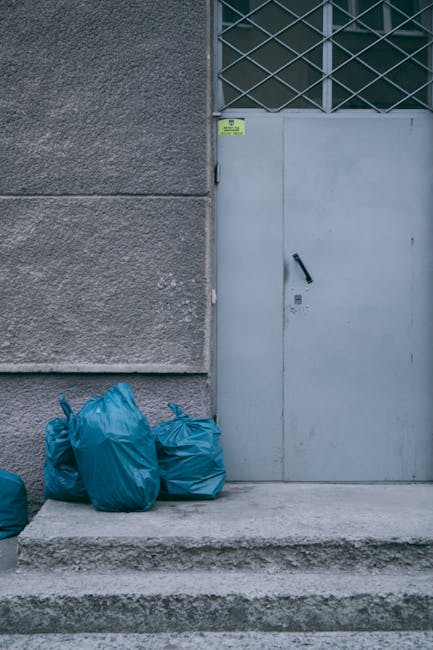 A narrow outdoor alleyway adjacent to a residential building, featuring a large grey plastic skip filled with black and white rubbish bags and various waste materials, positioned on the left side. Sev